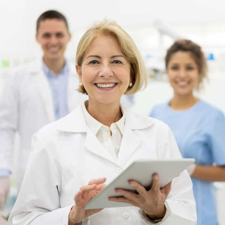 Woman in a white coat holding a tablet with two colleagues in the background.